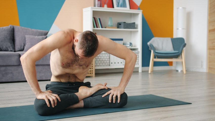 Topless guy is execising abdomen muscles practising yogic breathing technique at home sitting on yoga mat moving belly. People and healthy lifestyle concept.